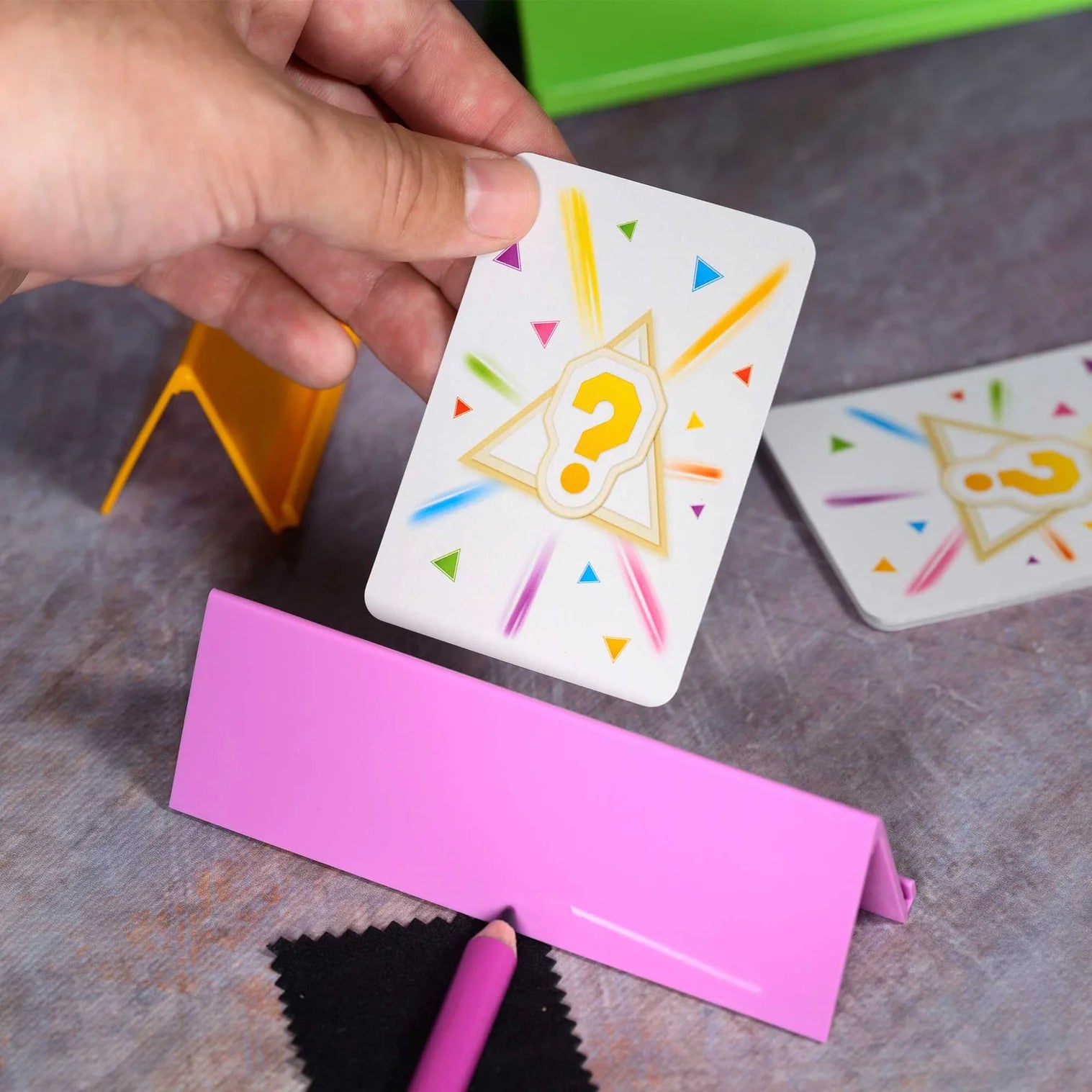 Hand holding a card with a colorful star and question mark on a gray surface with another card and pink strip.