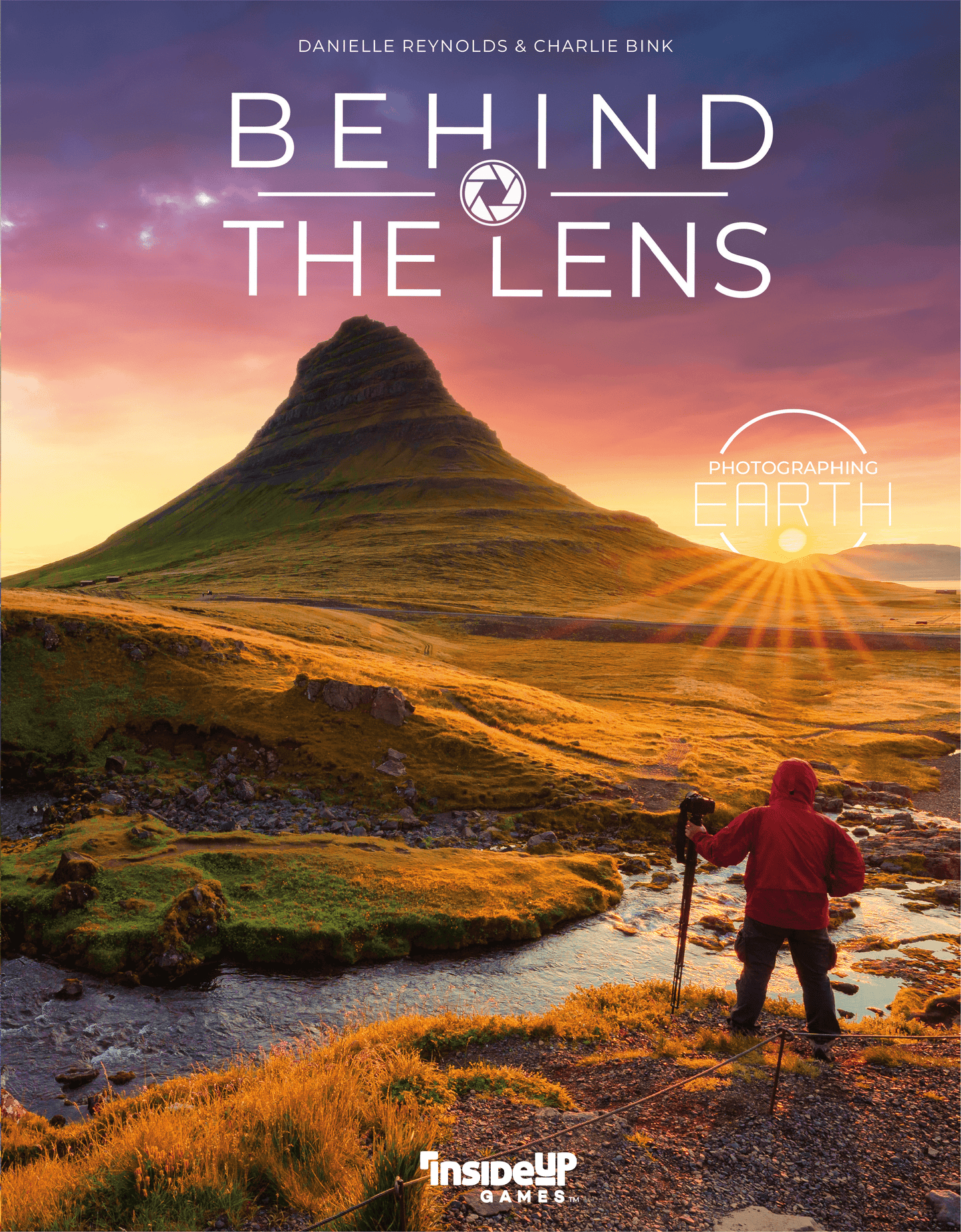 Book cover for 'Behind the Lens: Photographing Earth' by Danielle Reynolds & Charlie Bink, featuring a dramatic mountain landscape at sunset with a hiker in a red jacket standing by a stream in the foreground.
