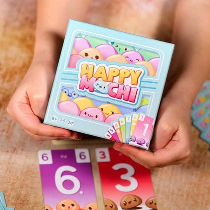 Person holding a 'Happy Mochi' card game box with colorful illustrations on a wooden surface.