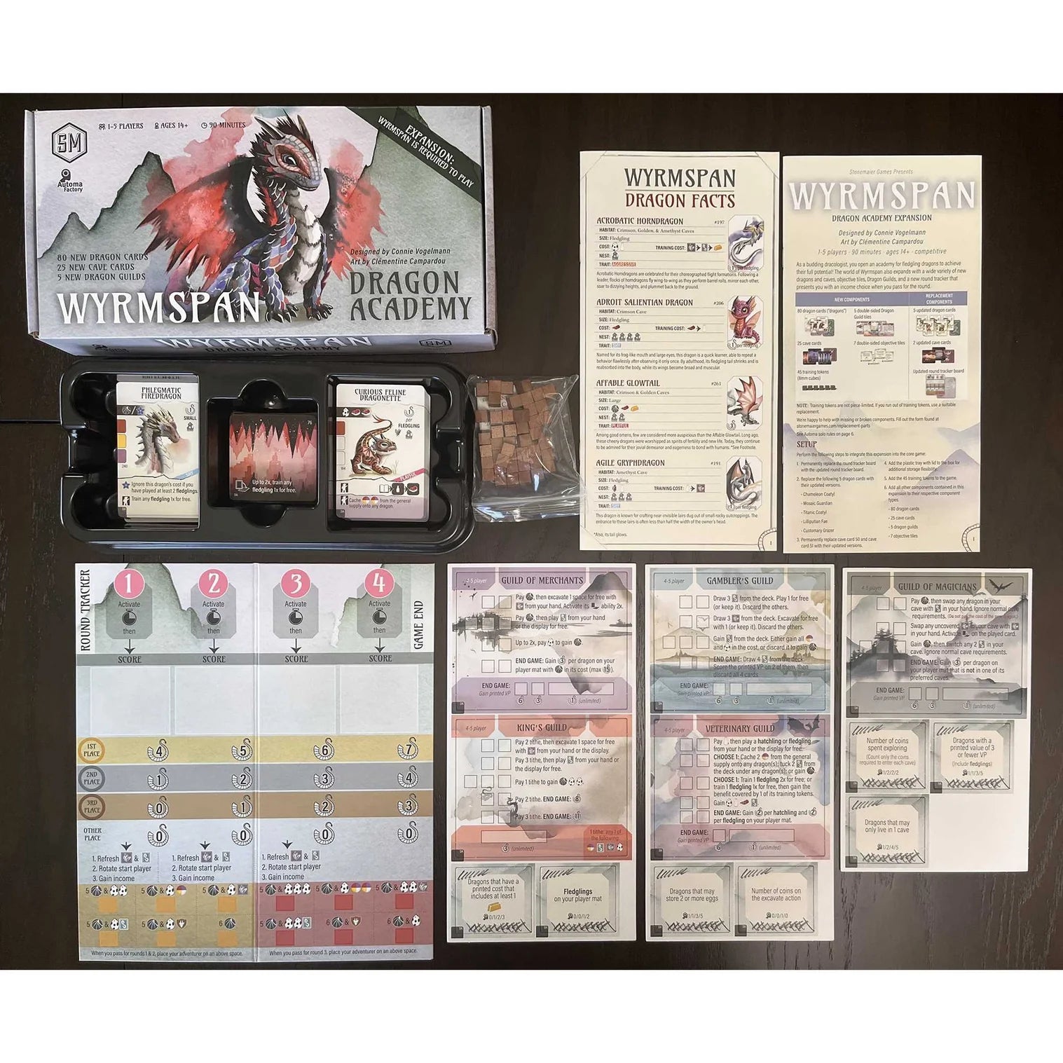 Wyrmspan board game components including cards, dragon academy box, and fact sheets on a dark surface.